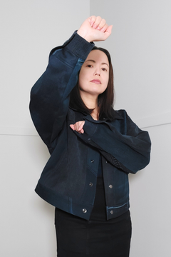 A woman wearing a cropped dark navy denim jacket over a black top and black skirt, posing with one arm raised against a light gray background. 