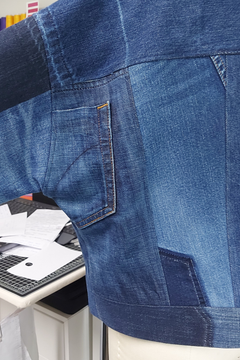 A close-up of a blue denim jacket with patchwork panels and contrast stitching, shown on a mannequin torso in an indoor setting.