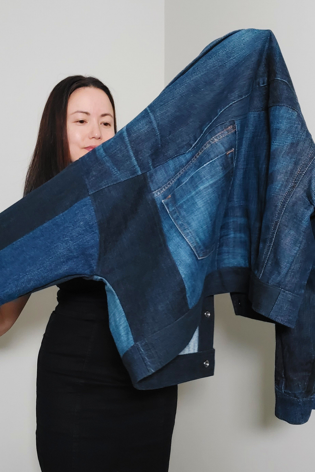 A woman holds up a cropped patchwork denim jacket made from various shades of blue denim, including light blue and dark indigo, against a plain light gray wall. Upcycled denim jacket by September Archive. Photographed by Sheila Wong.