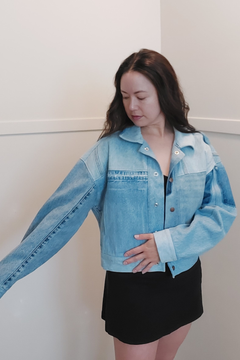 A woman wearing a light blue cropped denim jacket with contrast stitching and a black mini skirt, standing against a plain off-white wall. Upcycled denim jacket by September Archive. Photo by Sheila Wong. 