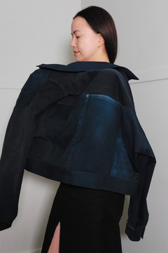A woman wearing a cropped dark denim jacket with a large collar and patchwork back panels, shown from the back against a plain light gray wall. Upcycled denim jacket by September Archive. Photographed by Sheila Wong.