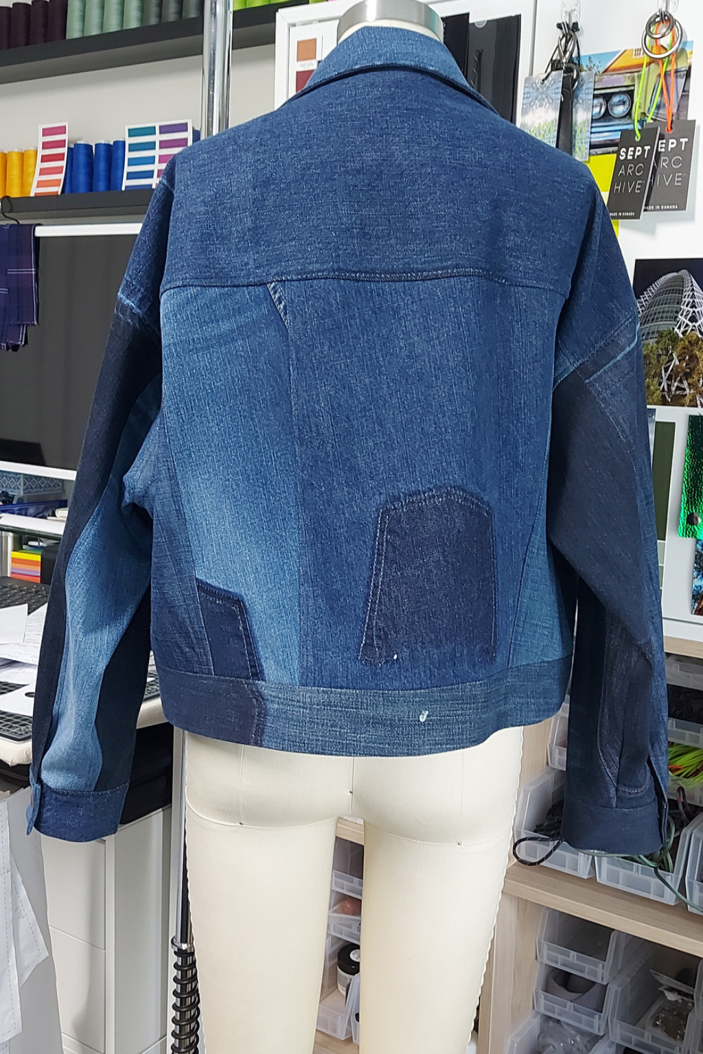 A blue denim jacket with contrasting dark blue denim sleeves and back patch pockets, displayed on a cream-colored mannequin torso in a retail or workshop setting.