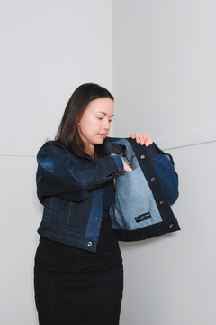 A woman wearing a dark blue patchwork denim jacket and black skirt, holding the jacket open to show the light blue denim lining. The jacket features a dark blue denim front and sleeves with a contrasting light blue denim lining. Upcycled denim jacket by September Archive. Photographed by Sheila Wong. 