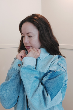A woman with long dark brown hair wearing a light blue denim jacket, looking down and adjusting the collar with her hands. Upcycled denim jacket by September Archive. Photo by Sheila Wong. 