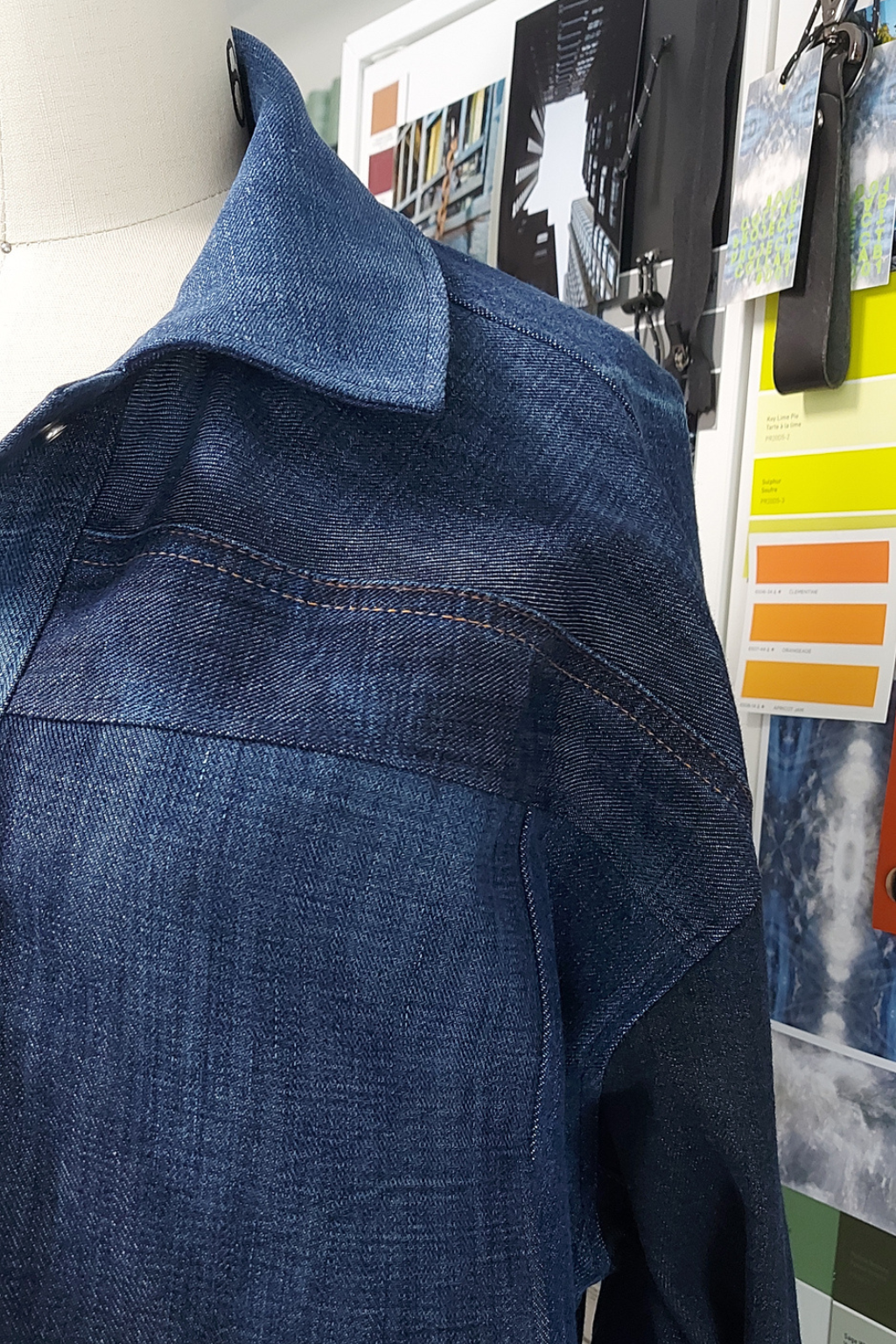 A close-up of a blue denim shirt or jacket displayed on a mannequin torso, with a collar and textured fabric visible against an indoor wall with posters and color cards.