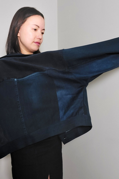 A woman wearing a dark navy blue patchwork jacket with wide sleeves, shown from the side against a plain light gray wall. Upcycled denim jacket by September Archive. Photographed by Sheila Wong.