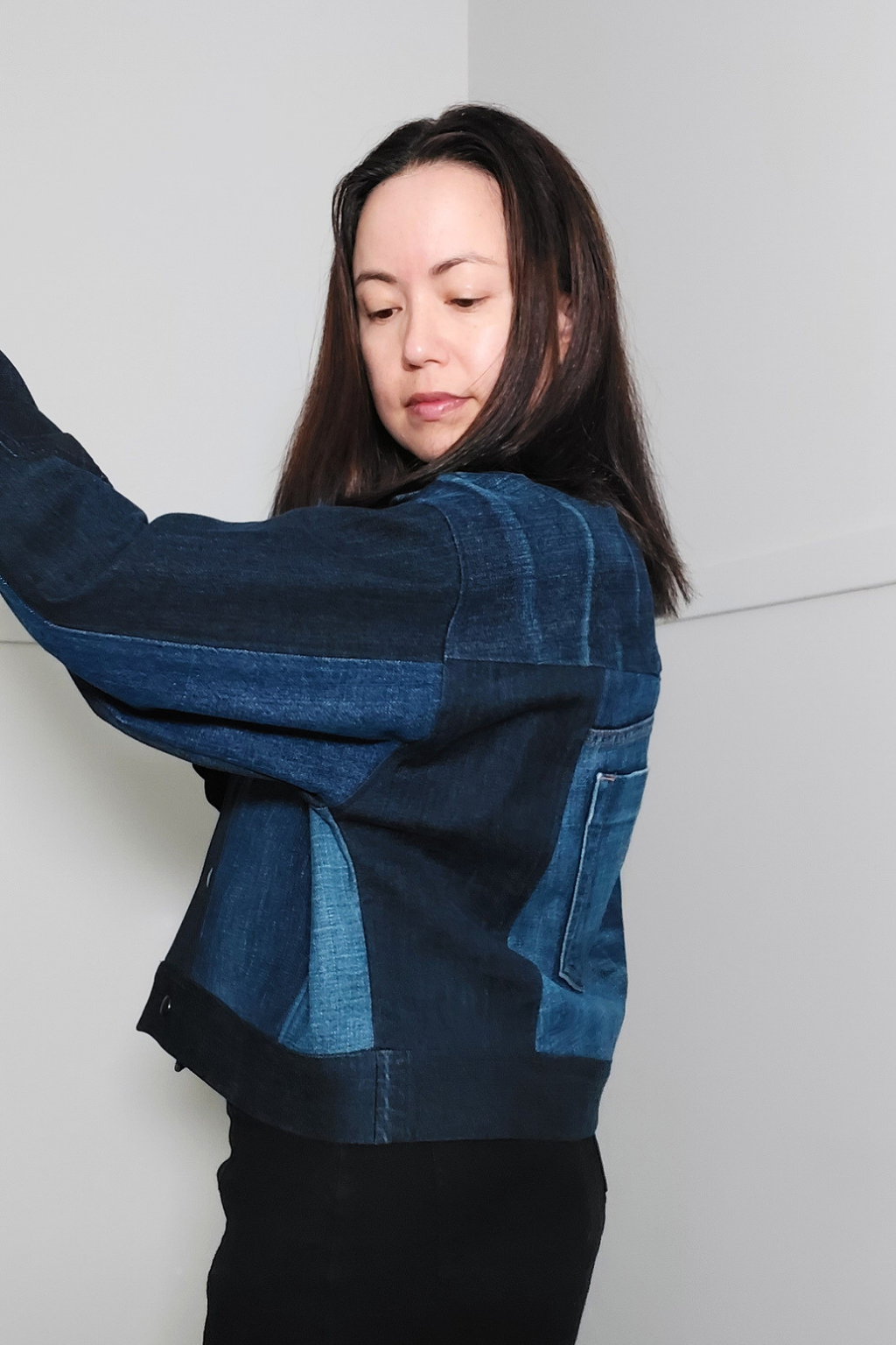 A woman wearing a dark blue patchwork denim jacket, posing with one arm raised against a plain light gray wall. Upcycled denim jacket by September Archive. Photographed by Sheila Wong.