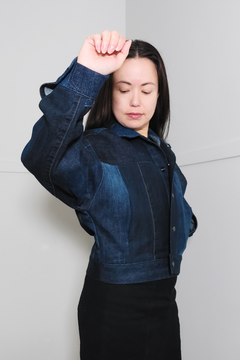 A woman wearing a cropped dark blue denim jacket with contrasting lighter blue denim panels on the chest and sleeves, paired with a black skirt, posing against a light gray wall. Upcycled denim jacket by September Archive. Photographed by Sheila Wong.