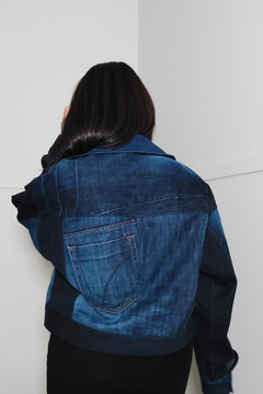 A person viewed from behind wearing a cropped denim jacket with contrasting dark navy sleeves and a back pocket, standing against a plain white wall. Upcycled denim jacket by September Archive. Photographed by Sheila Wong.