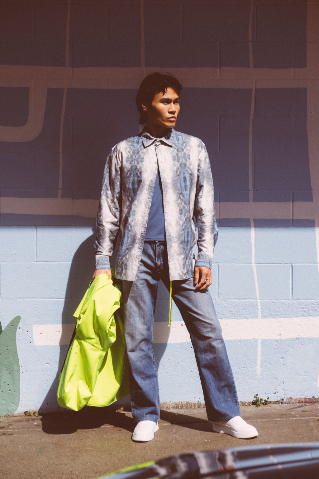 A man stands outdoors against a painted brick wall, wearing a light blue and white patterned overshirt over a blue shirt, blue jeans, and white sneakers, while holding a bright neon yellow-green jacket. Collaboration print 001 tailored shirt by September Archive. Captured by Trevor Brady.