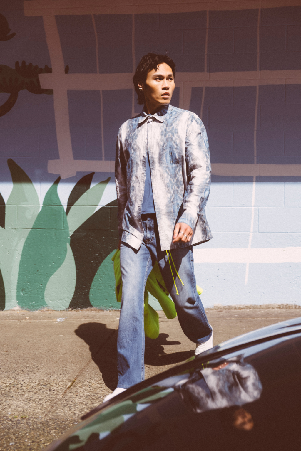 A man wearing a light blue and white patterned button-up shirt, blue jeans, and white sneakers walks outdoors in front of a painted wall with green leaf-like graphics. Collaboration print 001 tailored shirt by September Archive. Captured by Trevor Brady.
