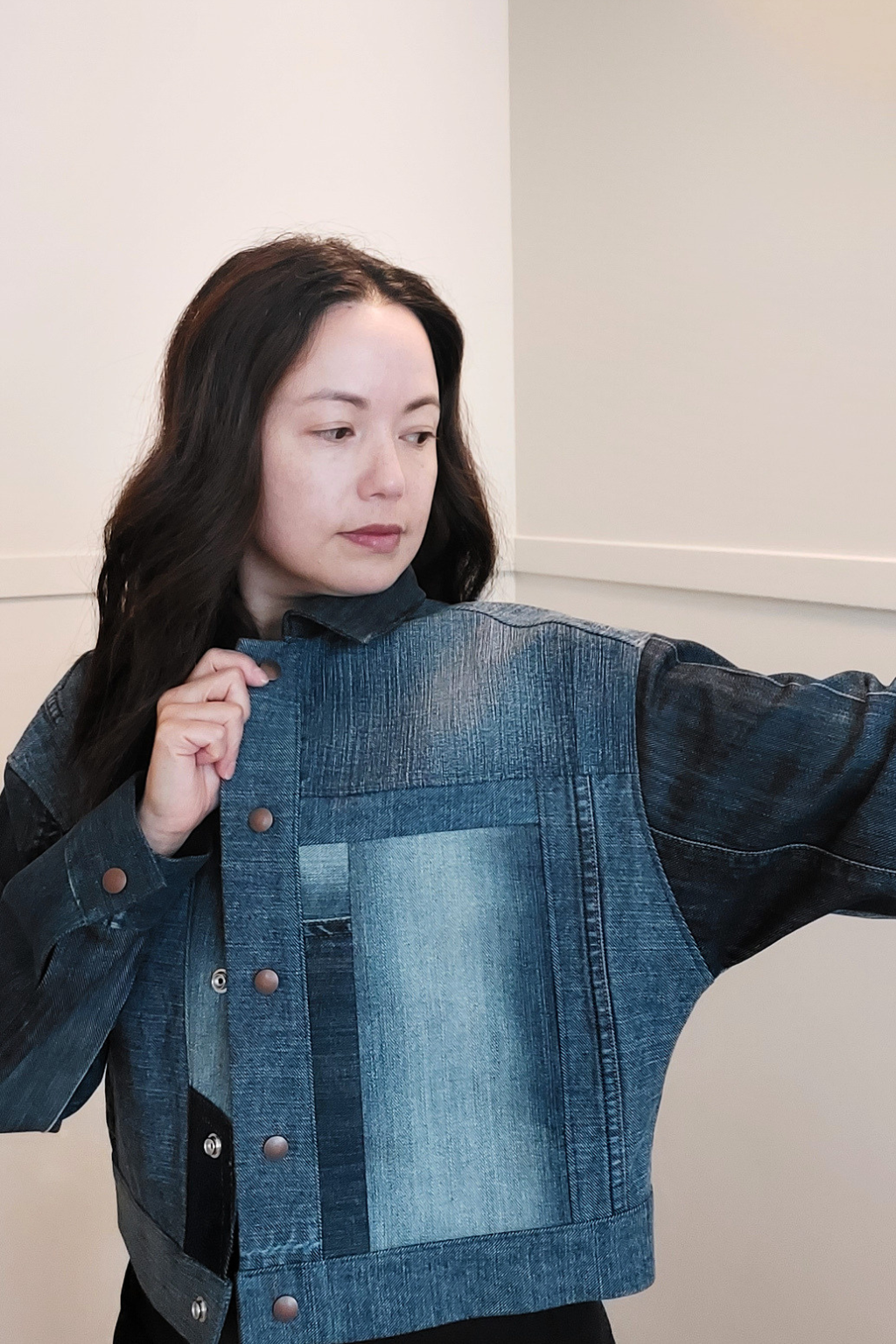 A woman wearing a cropped denim jacket with a patchwork design, featuring a light blue denim front panel, dark blue denim sleeves, and copper-toned buttons, set against a plain indoor wall. Upcycled denim jacket by September Archive. Photo by Sheila Wong. 