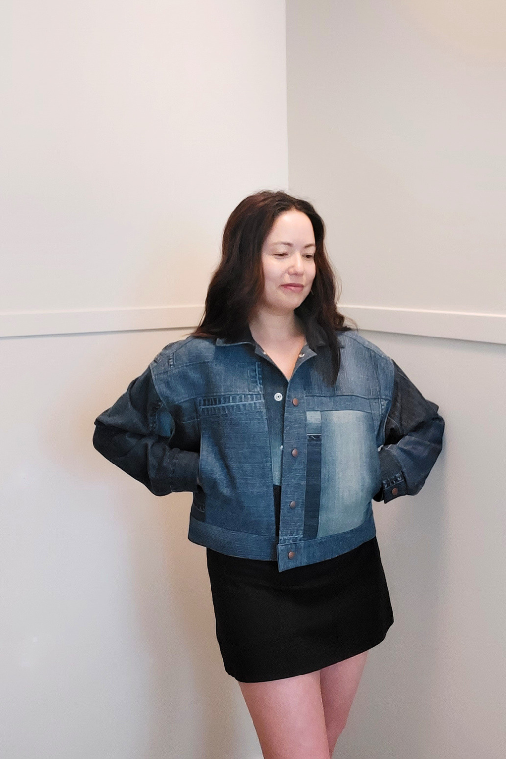 A woman modeling a cropped denim jacket with dark blue patchwork panels and black leather sleeves, paired with a black mini skirt, standing against a plain off-white wall. Upcycled denim jacket by September Archive. Photo by Sheila Wong. 