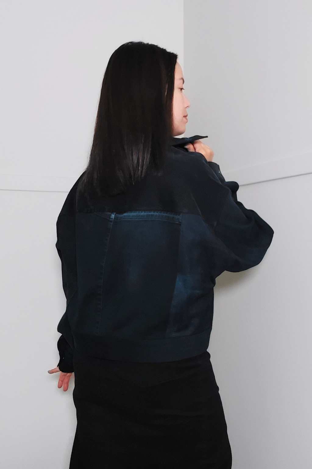 A woman with long black hair is shown from the back wearing a dark denim jacket with a patchwork design of dark blue and black denim panels, paired with a black skirt, set against a minimalist light gray corner wall. Upcycled denim jacket by September Archive. Photographed by Sheila Wong.