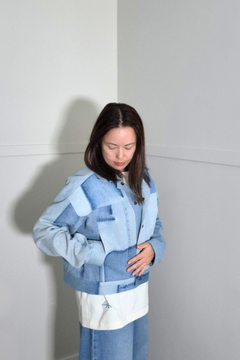 A woman wearing a light blue denim patchwork jacket with a white panel at the hem, standing in a minimalist gray room corner. Upcycled denim jacket by September Archive Apparel. Canadian made denim jacket. One of a kind cropped denim jacket made in Vancouver, Canada. Photographed by Sheila Wong. 