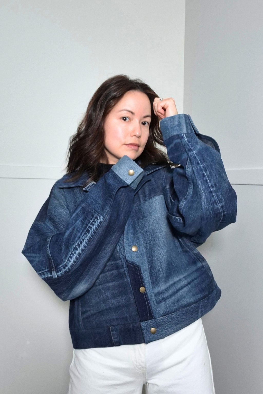 A woman wearing a cropped, oversized dark blue denim jacket with unique patchwork seams and gold-tone snap buttons, paired with white pants, posing against a light gray wall. Upcycled denim jacket by September Archive Apparel. Canadian made denim jacket. One of a kind cropped denim jacket made in Vancouver, Canada. Photographed by Sheila Wong. 