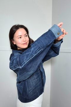 A woman wearing a dark blue denim jacket with unique contrast stitching and buttoned cuffs, posing with her arms raised against a plain light gray background. Upcycled denim jacket by September Archive Apparel. Canadian made denim jacket. One of a kind cropped denim jacket made in Vancouver, Canada. Photographed by Sheila Wong. 