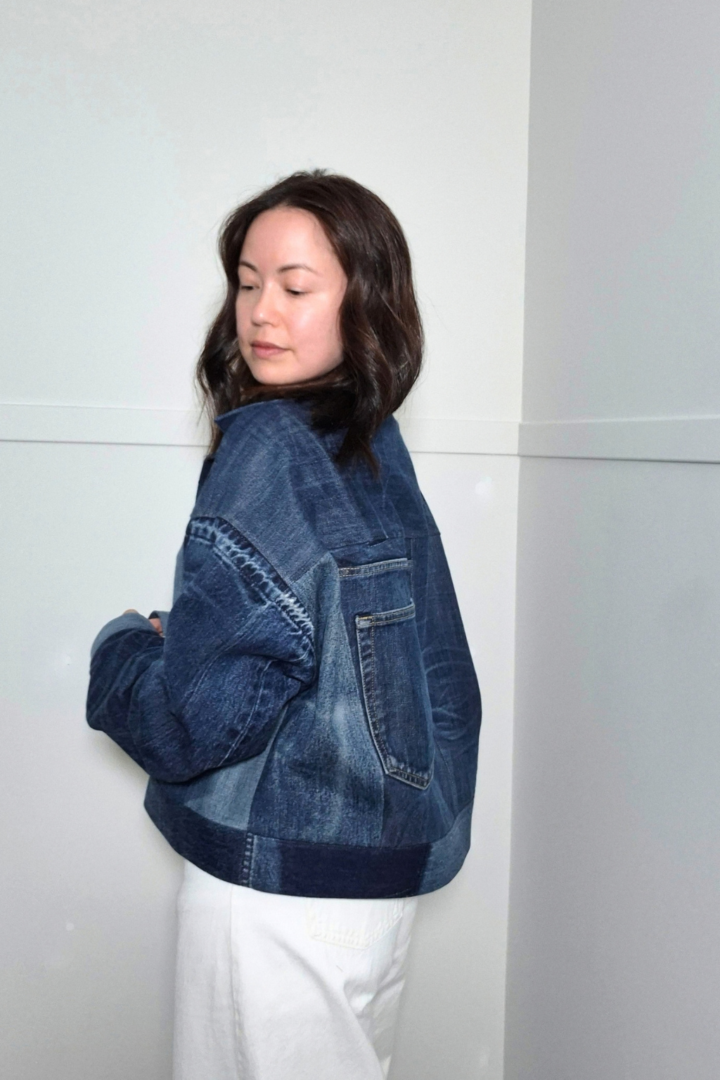 A woman wearing a cropped, patchwork-style blue denim jacket with a high collar and front patch pocket, paired with white pants, standing against a minimalist light gray wall. Upcycled denim jacket by September Archive Apparel. Canadian made denim jacket. One of a kind cropped denim jacket made in Vancouver, Canada. Photographed by Sheila Wong. 