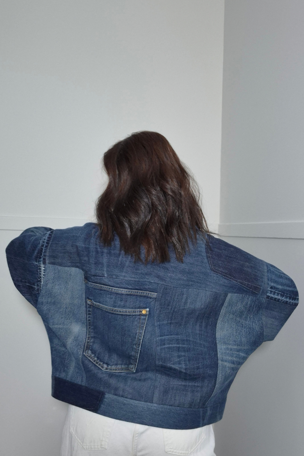 A person viewed from behind wearing a cropped denim jacket with patchwork panels and a large back pocket, paired with white pants, standing against a plain light gray wall. Upcycled denim jacket by September Archive Apparel. Canadian made denim jacket. One of a kind cropped denim jacket made in Vancouver, Canada. Photographed by Sheila Wong. 