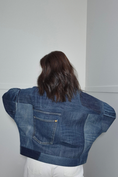 A person viewed from behind wearing a cropped denim jacket with patchwork panels and a large back pocket, paired with white pants, standing against a plain light gray wall. Upcycled denim jacket by September Archive Apparel. Canadian made denim jacket. One of a kind cropped denim jacket made in Vancouver, Canada. Photographed by Sheila Wong. 